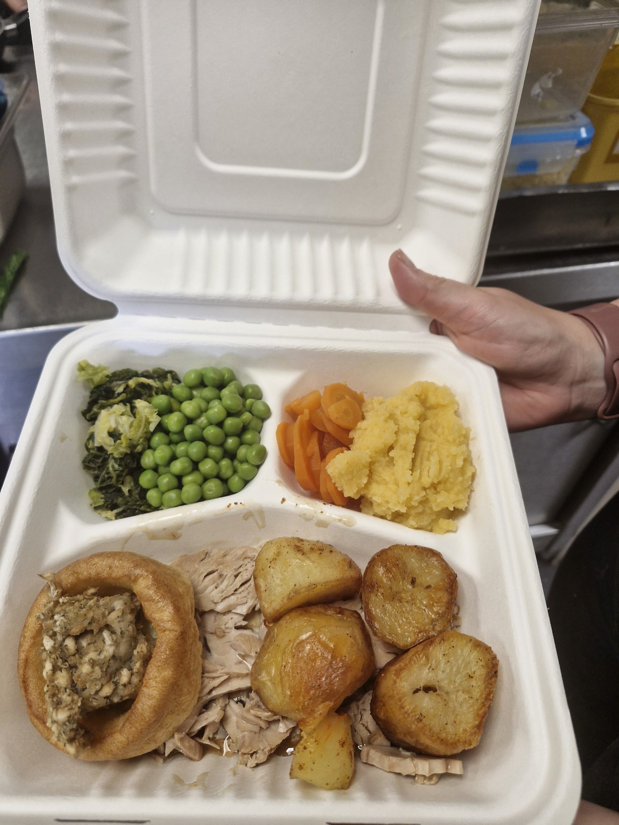 A roast dinner packed in a takeaway box: roast potatoes, a Yorkshire pudding with stuffing, peas, carrots, swede and greens